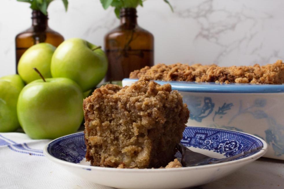 Apple cinnamon crumb cake is sliced and served on a blue china saucer. Apples, amber bottles of flowers, and the casserole dish with more cake is directly behind it.