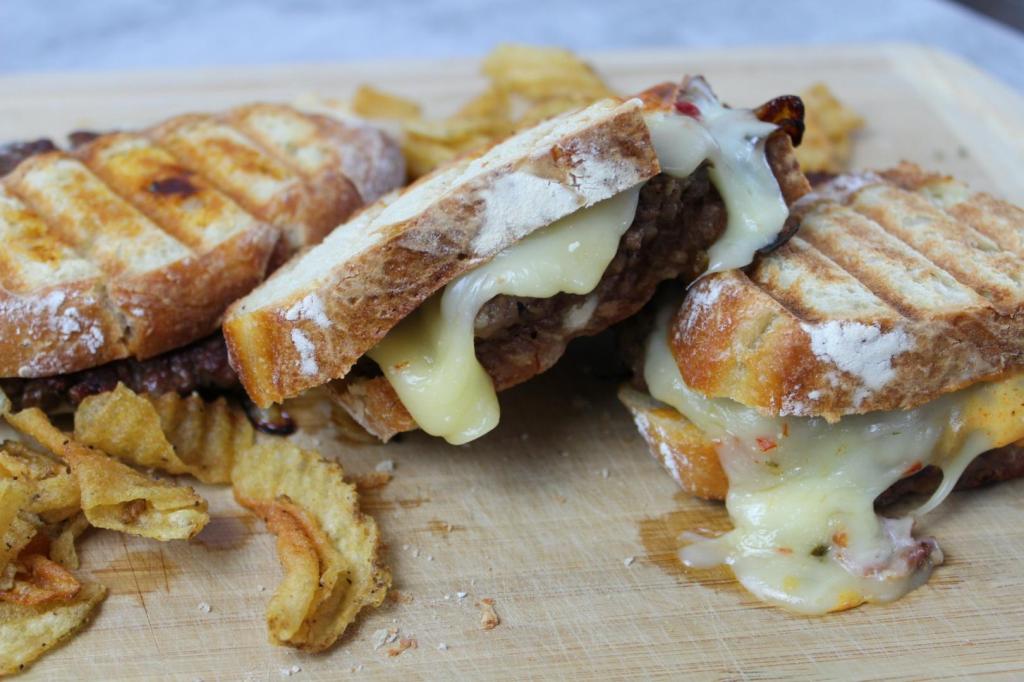 The Best Patty Melt recipe consists of three patty melt burgers toasted to perfection on a cutting board, surrounded by kettle potato chips.