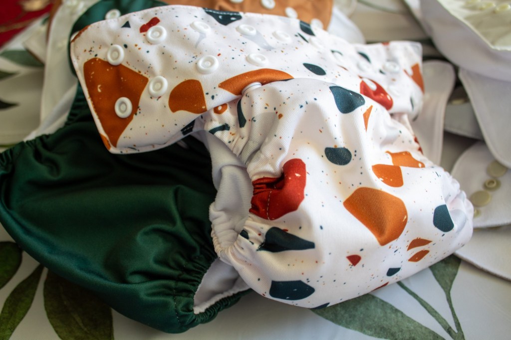 Noras Nursery cloth diapers are shown in a pile for a review. A green and multicolored one are shown.