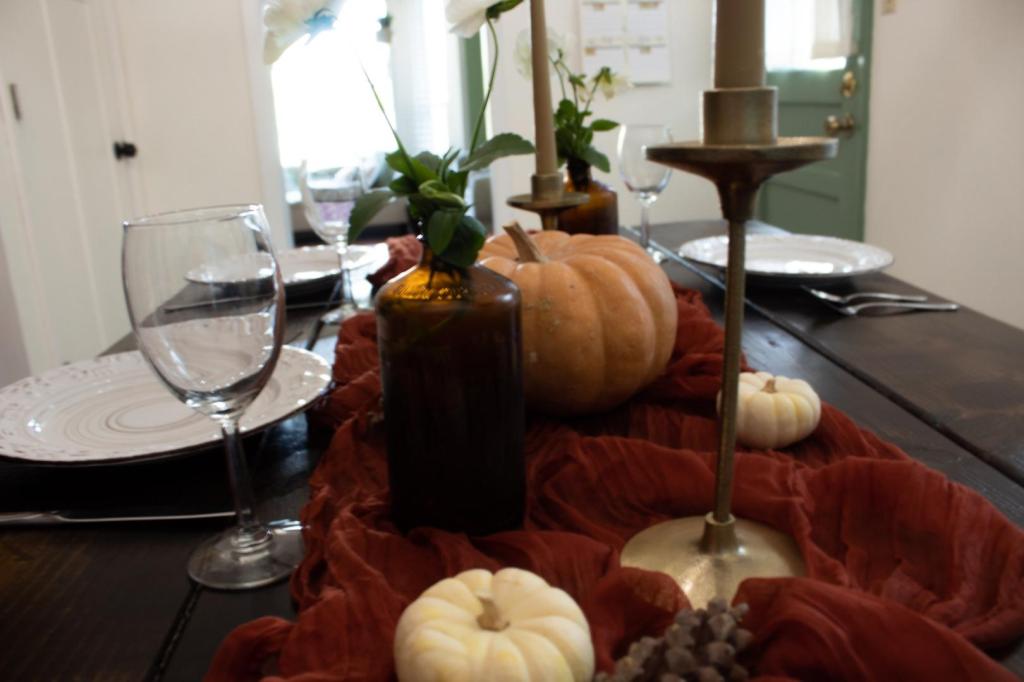 A fall table setting (autumn tablescape) is decorated with a terra cotta table runner, mini white pumpkins, wine glasses, a large pumpkin centerpiece, candles, and amber bottles filled with delicate white flowers.
