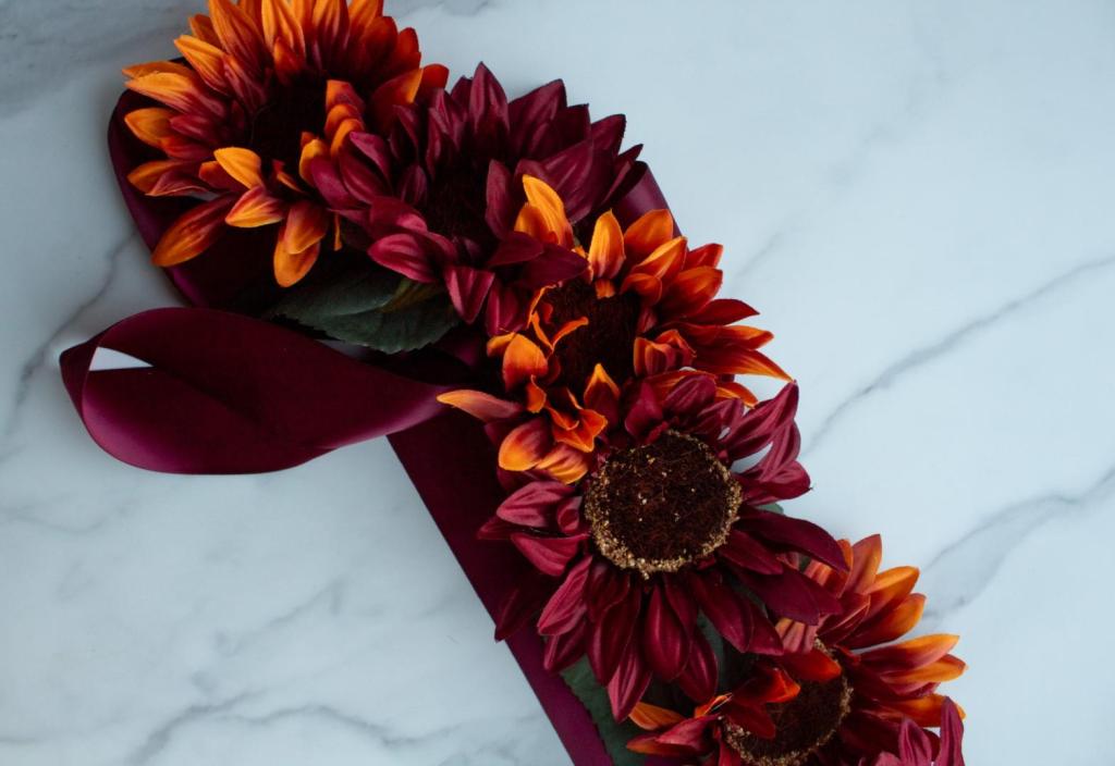 A maternity sash belt lays on a countertop and is burgundy with lots of fall sunflowers.