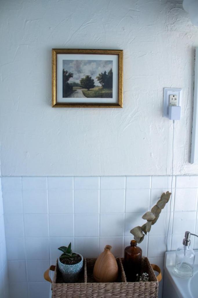 A Miss Mustard Seed landscape art print sits above a toilet in a small bathroom.