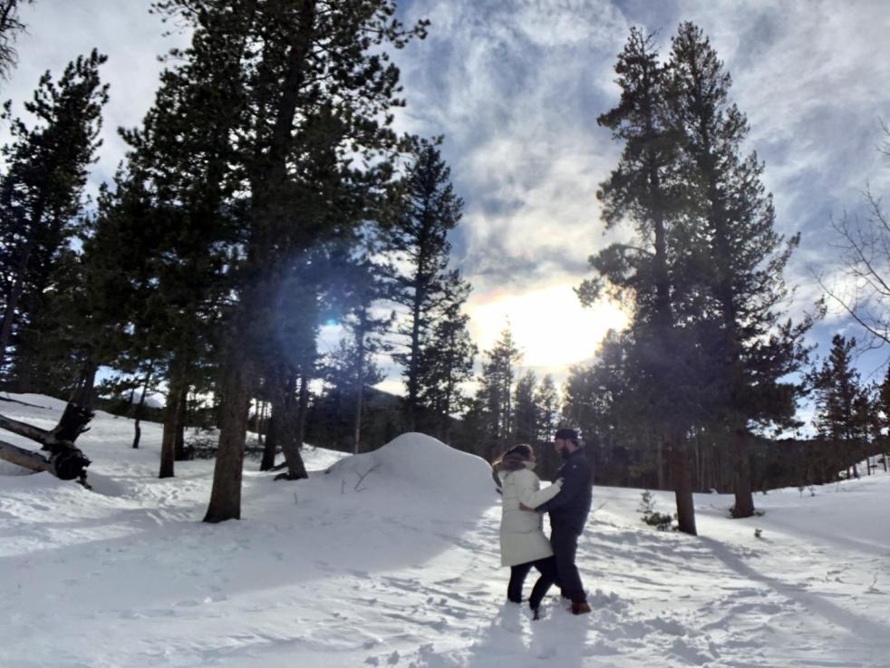 New family traditions consists of a man and woman embracing on a mountains near snowy pine trees.