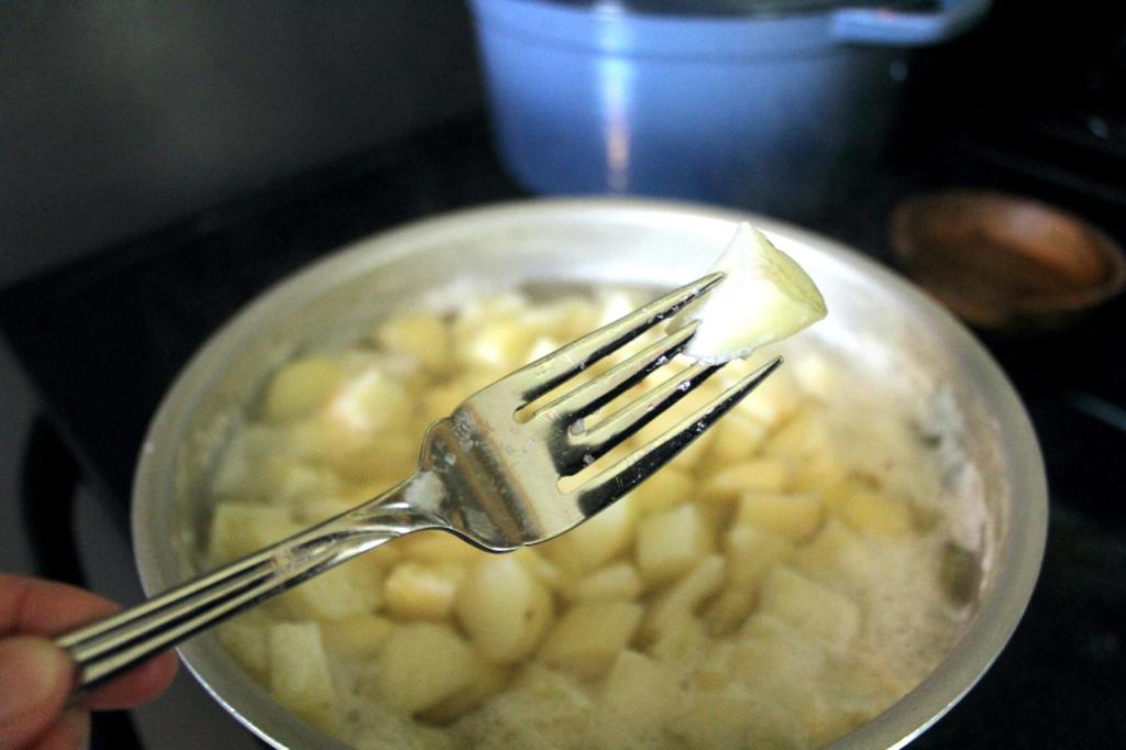 With a pot of boiling potatoes in the background, a woman tests the texture of a piece using a fork.