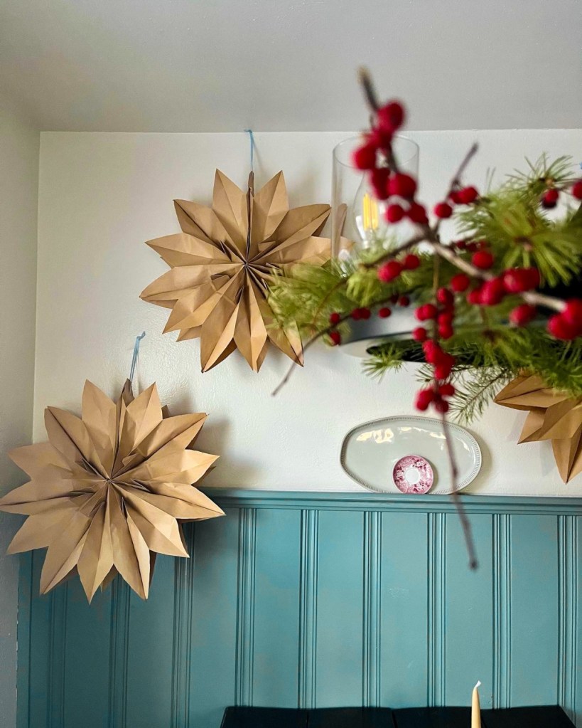 A paper snowflake tutorial is followed to create paper snowflakes that hang in a kitchen behind a chandelier during Christmas.