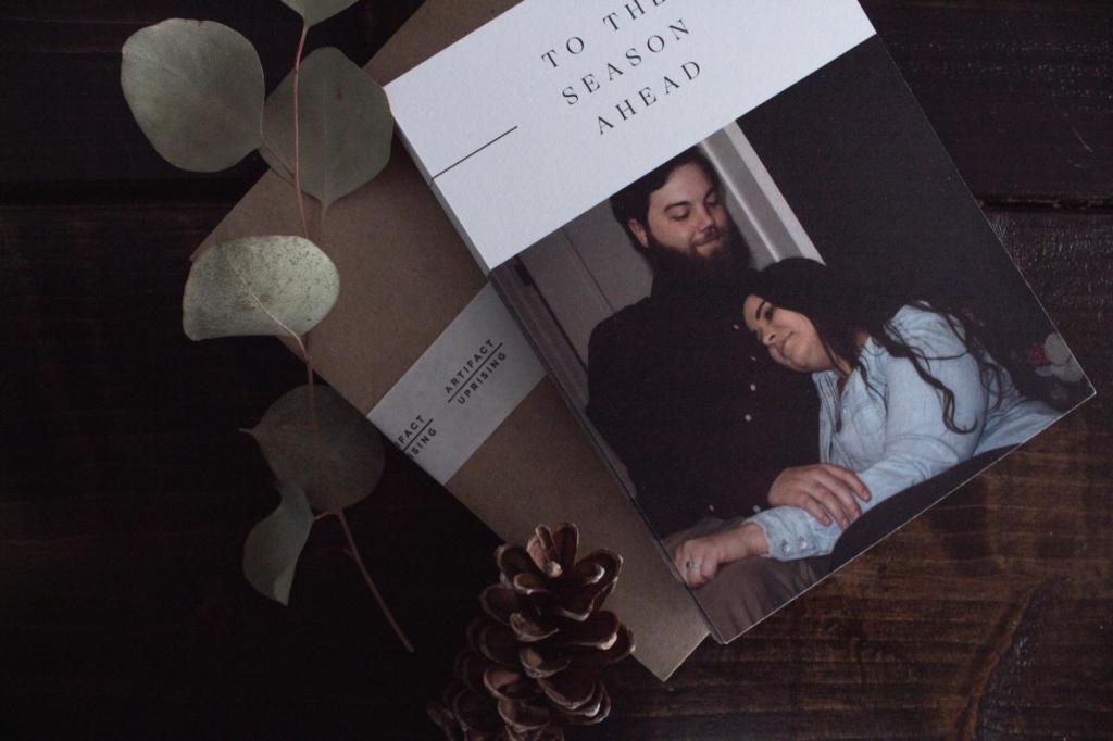 A man and woman embrace on a Christmas card laid on a tabletop next to pine cones.