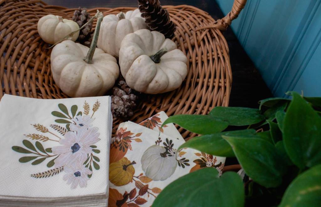 A small centerpiece includes a basket full of pumpkins, festive napkins, and a plant.