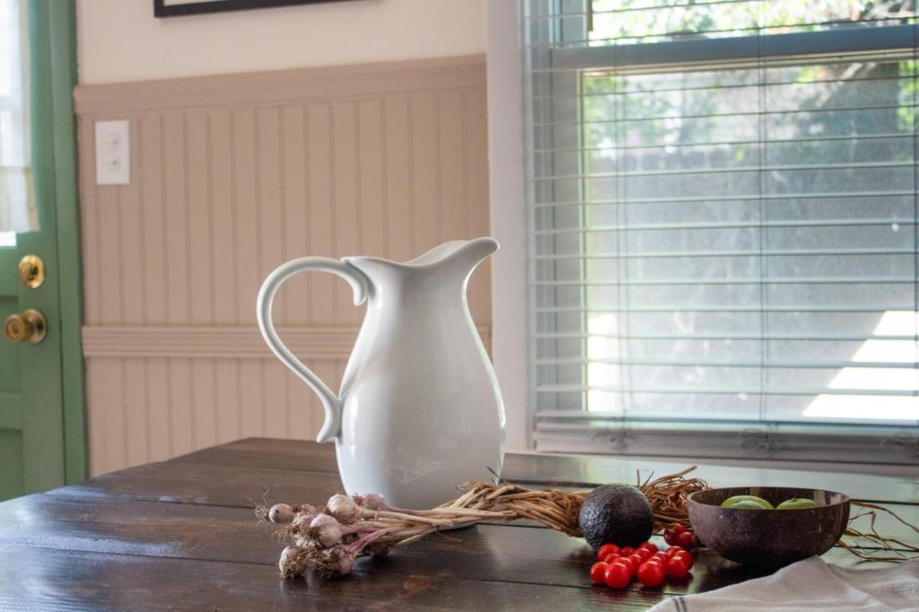 A farmhouse table has produce from a garden and a porcelain pitcher on it in a cottagecore kitchen.