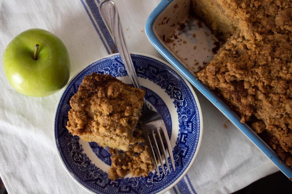 Apple cinnamon cake is sliced from a casserole dish and served next to an apple on a blue china saucer.