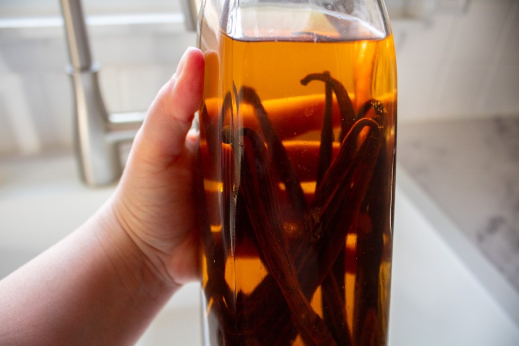 A vanilla extract recipe uses vodka and vanilla pods which are shown in a bottle as a woman holds it up in a white kitchen.