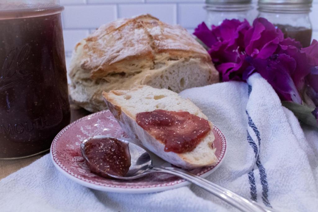 Toast is smothered in a fig and tomato jam recipe over a saucer and linen with a jar of jam and purple flowers nearby, along with a loaf of bread.
