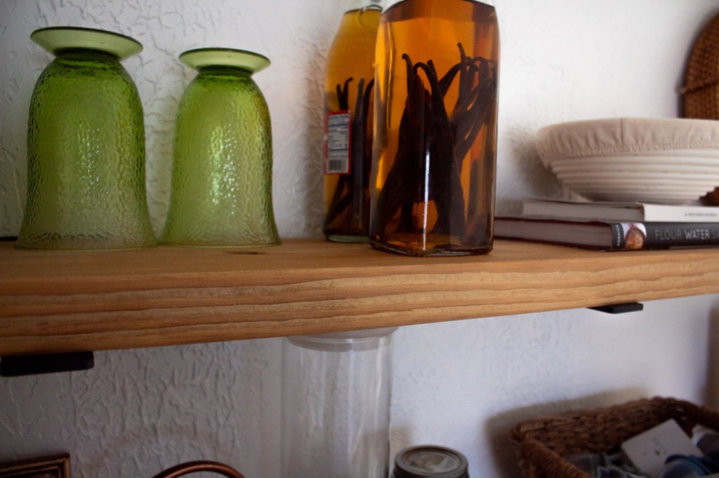 Pure vanilla extract sits bottled on the top shelf of a kitchen open shelves.